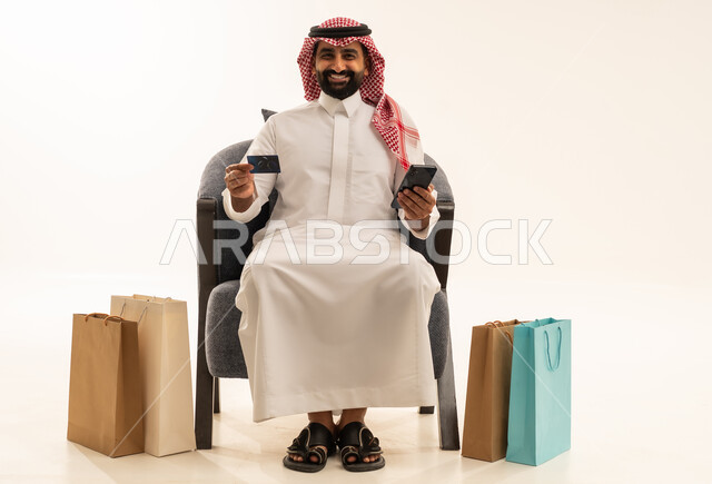 الشراء والدفع عن طريق بطاقة الإئتمان البنكية، إتمام عملية الطلبات باستخدام الهاتف المحمول، مفهوم التسوق عن بعد, بورتريه لرجل عربي خليجي سعودي يرتدي الثوب التقليدي والشماغيجلس على كرسي بجانبة أكياس ورقية ملونة, التبضع والاستفادة من العروض والتخفيضات, خلفية بيضاء