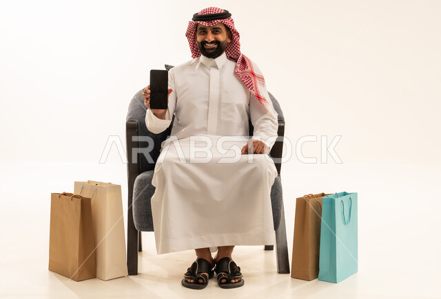 Buying products and items online, a portrait of a Saudi Arabian Gulf man wearing traditional attire and a ghutrah sitting on a chair holding a mobile phone in his hand with paper bags beside him, displaying a blank black screen on the mobile, white background.