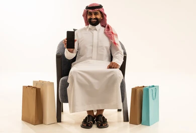 شراء المنتجات و الأغراض عبر الانترنت , بورتريه لرجل عربي خليجي سعودي يرتدي الثوب التقليدي و الشماغ يجلس على الكرسي يمسك الهاتف المحمول بيده و بجانبه أكياس ورقية , عرض شاشة سوداء فارغة عبر الجوال ، خلفية بيضاء