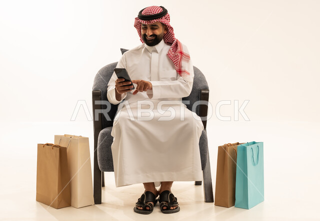 ايماءات السعادة و السرور، تصفح مواقع التواصل الاجتماعي، مفهوم التسوق الالكتروني  , بورتريه لرجل عربي خليجي سعودي يرتدي الثوب التقليدي و الشماغ يجلس على الكرسي يمسك الهاتف المحمول بيده و بجانبه أكياس ورقية , خلفية بيضاء
