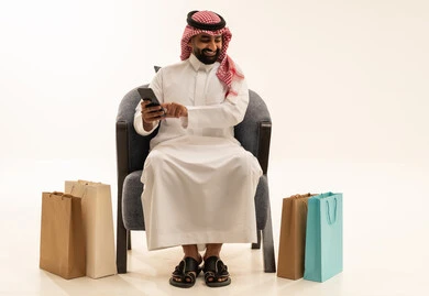 مفهوم التسوق الالكتروني ,تصفح مواقع التواصل الاجتماعي , بورتريه لرجل عربي خليجي سعودي يرتدي الثوب التقليدي و الشماغ يجلس على الكرسي يمسك الهاتف المحمول بيده و بجانبه أكياس ورقية , خلفية بيضاء
