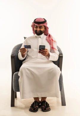Online shopping, the concept of remote payment and settlement through mobile applications, the use of electronic bank cards, a portrait of a Saudi Gulf Arab man wearing a white thobe and a ghutrah sitting on a chair holding a mobile phone, white background.