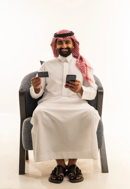 Online shopping, the concept of remote payment and settlement through mobile applications, the use of electronic bank cards, a portrait of a Saudi Gulf Arab man wearing a white thobe and a ghutrah sitting on a chair holding a mobile phone, white background.