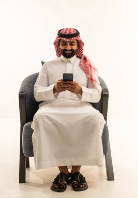 استخدام الأجهزة التقنية الحديثة للتسلية و الترفيه، تصفح وسائل التواصل الاجتماعي  , بورتريه لرجل عربي خليجي سعودي يرتدي الثوب الابيض و الشماغ يجلس على كرسي يمسك الهاتف المحمول بيده , خلفية بيضاء