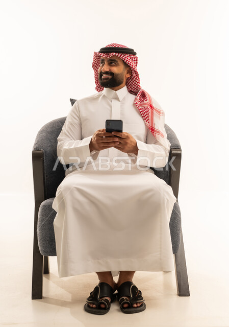 Looking at something, using modern technological devices for entertainment and leisure, browsing social media, a portrait of a Saudi Arabian Gulf man wearing a white thobe and a ghutrah sitting on a chair holding a mobile phone in his hand, white background.