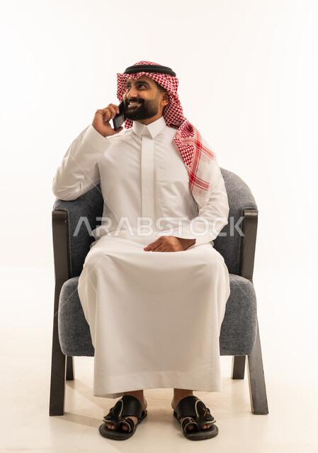 تبادل الأحاديث مع الأهل والأصدقاء، إجراء مكالمة هاتفية باستخدام الهاتف المحمول، بورتريه لرجل عربي خليجي سعودي يرتدي الثوب الابيض و الشماغ يجلس على كرسي يقوم بالتحدث بهاتفه المحمول بتعابير الفرح والسرور، خلفية بيضاء
