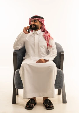 تبادل الأحاديث مع الأهل والأصدقاء، إجراء مكالمة هاتفية باستخدام الهاتف المحمول، بورتريه لرجل عربي خليجي سعودي يرتدي الثوب الابيض و الشماغ يجلس على كرسي يقوم بالتحدث بهاتفه المحمول بتعابير الفرح والسرور، خلفية بيضاء