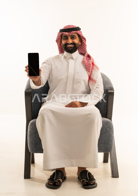 The concept of technological and technical developments, a portrait of a Gulf Arab Saudi man wearing a white thobe and a shemagh sitting on a chair displaying a blank black screen via a mobile phone, keeping up with modern contemporary progress, white background.