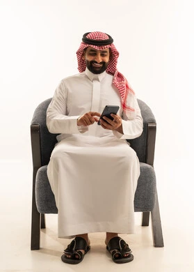 تصفح وسائل التواصل الاجتماعي , استخدام الأجهزة التقنية الحديثة للتسلية و الترفيه , بورتريه لرجل عربي خليجي سعودي يرتدي الثوب الابيض و الشماغ يجلس على كرسي يمسك الهاتف المحمول بيده , خلفية بيضاء
