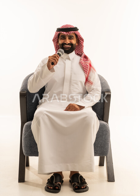 النظر للكاميرا بتعابير السرور، القاء الخطابات والكلمات باستخدام مكبر الصوت، القيام بالحوارات والنقاشات، بورتريه لرجل عربي خليجي عماني سعودي يرتدي الثوب الابيض و الشماغ يجلس على كرسي يحمل بيده مكبر الصوت، خلفية بيضاء