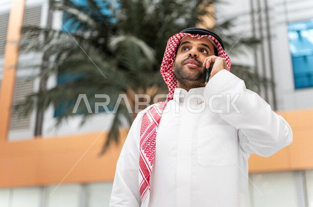 A Saudi Gulf man, making a phone call via a mobile phone inside the tourist hotel, using modern technical equipment, a working session, the concept of business management, making business deals, working remotely, e-marketing, a business trip, tourism and