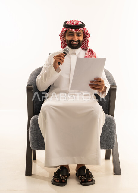 متابعة ومراجعة الخطط والمشاريع المكتبية، اعداد التقارير الخاصة بالعمل، تقديم الفقرات باستخدام الميكروفون، بورتريه لرجل عربي خليجي سعودي يرتدي الثوب التقليدي والشماغ يجلس على كرسي ينظر الى الاوراق البيضاء يمسك بيده المايك, التدقيق في قراءة الملاحظات، خلفية بيضاء