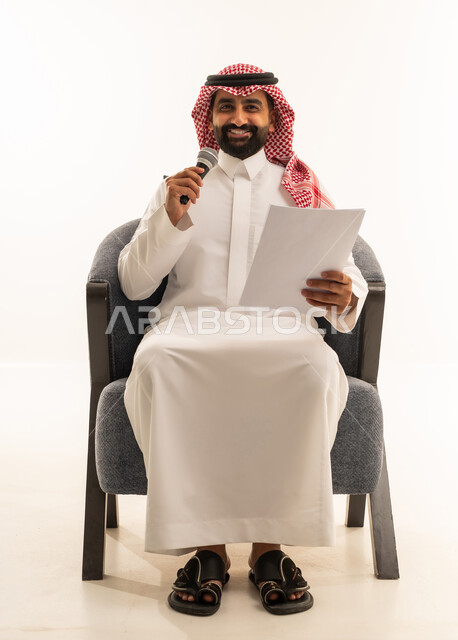 اعداد التقارير الخاصة بالعمل، تقديم الفقرات باستخدام الميكروفون ، متابعة ومراجعة الخطط والمشاريع المكتبية، بورتريه لرجل عربي خليجي سعودي يرتدي الثوب التقليدي والشماغ يجلس على كرسي ينظر الى الاوراق البيضاء يمسك بيده المايك, التدقيق في قراءة الملاحظات، خلفية بيضاء