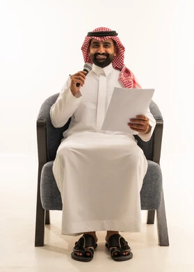 اعداد التقارير الخاصة بالعمل، تقديم الفقرات باستخدام الميكروفون ، متابعة ومراجعة الخطط والمشاريع المكتبية، بورتريه لرجل عربي خليجي سعودي يرتدي الثوب التقليدي والشماغ يجلس على كرسي ينظر الى الاوراق البيضاء يمسك بيده المايك, التدقيق في قراءة الملاحظات، خلفية بيضاء
