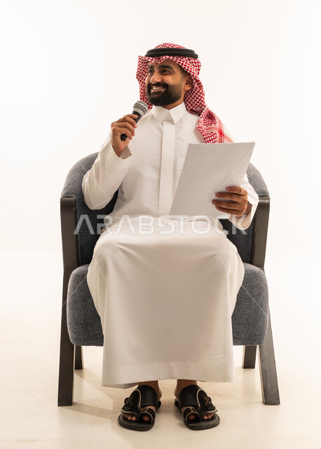 Presenting paragraphs using the microphone, preparing work-related reports, following up and reviewing office plans and projects, a portrait of a Saudi Gulf Arab man wearing traditional attire and a ghutrah sitting on a chair looking at white papers holding a microphone in his hand, proofreading notes, white background.