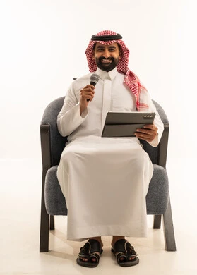 Artistic singing talents, recording a vocal clip, merging with the song, a portrait of a Saudi Gulf Arab man wearing traditional attire and a ghutrah sitting on a chair singing using a microphone while holding a tablet in his hands with expressions of enthusiasm, white background.