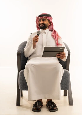 تسجيل مقطع صوتي غنائي، مواهب غنائية فنية، الاندماج مع الأغنية، بورترية لرجل عربى خليجى سعودى يرتدى الثوب التقليدى و الشماغ يجلس على الكرسى يقوم بالغناء باستخدام الميكروفون يمسك بيدية التابلت بتعابير الحماس، خلفية بيضاء