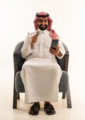 مواهب فنية في عالم الموسيقى، ممارسة هواية الغناء للتسلية والترفيه، الفن المسرحي الغنائي، تقديم العرض المسرحي، بورتريه لرجل عربي خليجي سعودي يرتدي الثوب و الشماغ يجلس على كرسي يحمل بيده الميكروفون و الهاتف المحمول ، خلفية بيضاء
