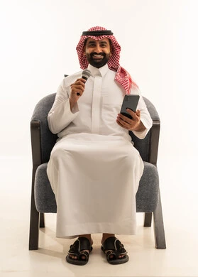 Looking at the camera with expressions of joy, practicing the hobby of singing for fun and entertainment, musical theater art, artistic talents in the world of music, presenting the theatrical performance, a portrait of a Saudi Arabian Gulf man wearing a thobe and a ghutrah sitting on a chair holding a microphone and a mobile phone, white background.