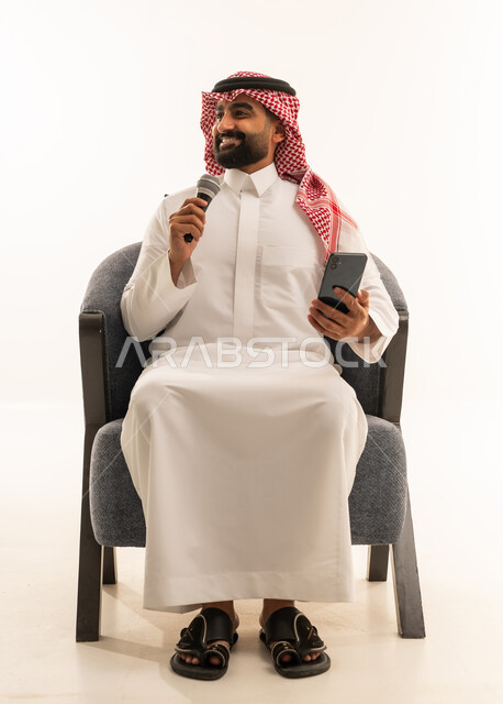 ممارسة هواية الغناء للتسلية والترفيه، الفن المسرحي الغنائي، مواهب فنية في عالم الموسيقى، تقديم العرض المسرحي، بورتريه لرجل عربي خليجي سعودي يرتدي الثوب و الشماغ يجلس على كرسي يحمل بيده الميكروفون و الهاتف المحمول ، خلفية بيضاء