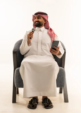 ممارسة هواية الغناء للتسلية والترفيه، الفن المسرحي الغنائي، مواهب فنية في عالم الموسيقى، تقديم العرض المسرحي، بورتريه لرجل عربي خليجي سعودي يرتدي الثوب و الشماغ يجلس على كرسي يحمل بيده الميكروفون و الهاتف المحمول ، خلفية بيضاء