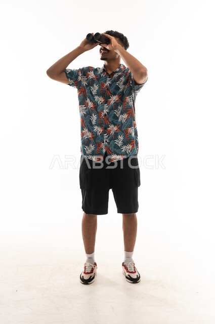 A Gulf Arab Omani man wearing casual attire is holding binoculars with both hands, looking up at something, full-body shot, portrait with a white background.