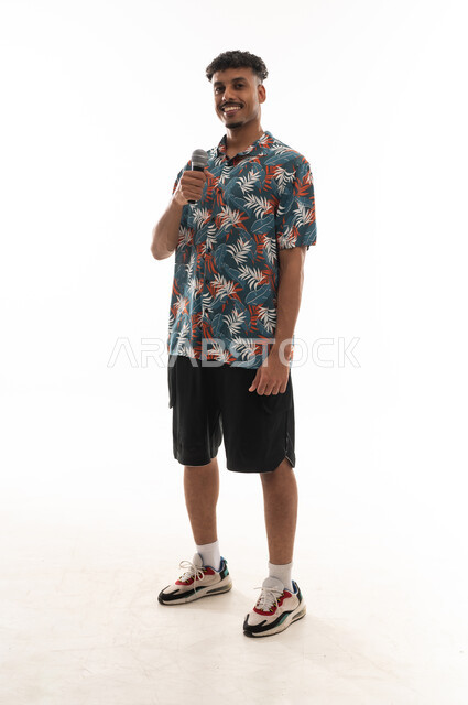 Looking at the camera while holding the microphone, an Arab Gulf Omani man wearing casual attire is singing into a loudspeaker, gestures of happiness, having a good time, full-body shot, portrait with a white background.