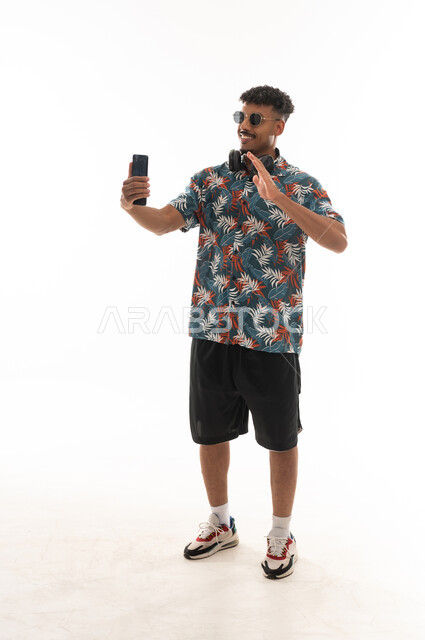 التلويح بكف اليد اشارة الترحيب و السلام , رجل عربي خليجي عماني يرتدي الزي الكاجوال يقوم باجراء مكالمة فيديو مباشرة , بورتريه خلفية بيضاء , الجسم بالطول الكامل
