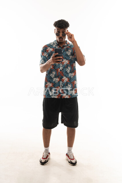 A Gulf Arab Omani man wearing casual attire, holding a mobile phone and raising sunglasses, with expressions of surprise and astonishment, portrait with a white background, full body.