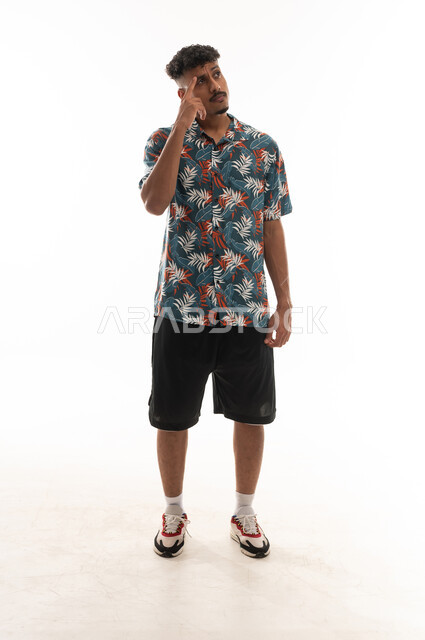 The index finger is placed on the forehead as a sign of deep thinking and confusion. An Arab Gulf Omani man is wearing casual attire, standing in front of the camera, looking up at something. Portrait with a white background, full-body shot.