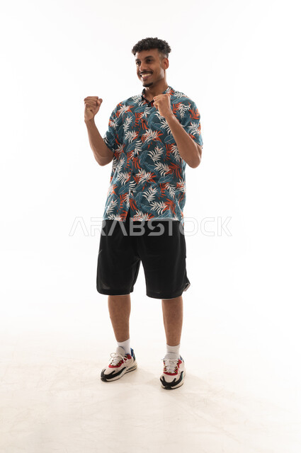 Clenching the fingers of both hands tightly and waving them in the air is a sign of victory and happiness. An Arab Gulf Omani man is wearing casual attire, with gestures of joy and delight, in a portrait against a white background, full-body.