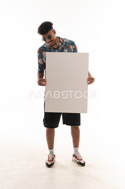 Looking happily at the large paper, an Arab Gulf Omani man wearing casual attire holds a blank sheet in front of the camera, portrait with a white background, full body.
