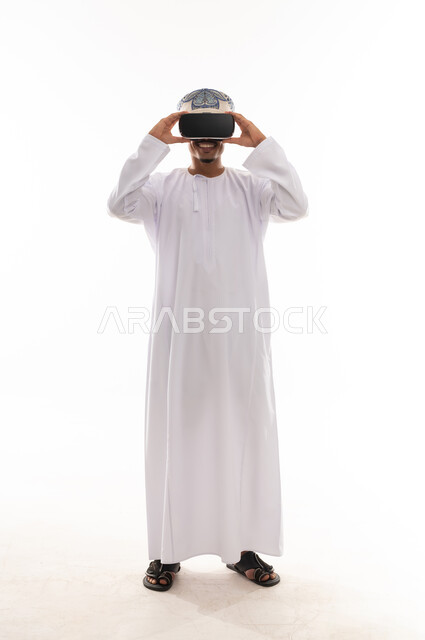 رجل عربي خليجي عماني يرتدي الدشداشة البيضاء و العمامة يمسك نظارة الواقع الافتراضي بيديه , ايماءات التشويق و الاثارة , الجسم بالطول الكامل , بورتريه خلفية بيضاء