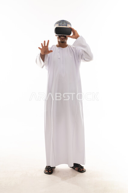نظارة محاكاة الواقع الحقيقي , رجل عربي خليجي عماني يرتدي الدشداشة البيضاء و العمامة يلبس نظارة الواقع الافتراضي , الشعور بالاثارة و التشويق , الجسم بالطول الكامل , بورتريه خلفية بيضاء