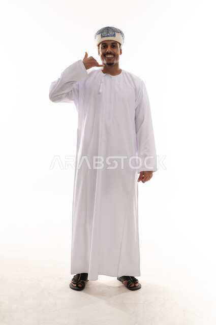 رفع اصبع الخنصر و الابهام اشارة الى طلب معاودة الاتصال , رجل عربي خليجي عماني يرتدي الدشداشة البيضاء و العمامة يقف امام الكاميرا , ايماءات السعادة و الابتسامة , الجسم بالطول الكامل , بورتريه خلفية بيضاء