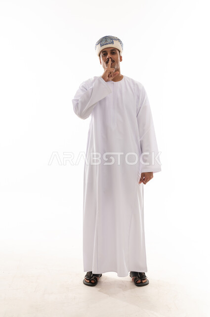 Placing the index finger on the mouth as a sign to request silence and stop talking, an Arab Gulf Omani man wearing a white dishdasha and turban, standing in front of the camera, full-body shot, portrait with a white background.