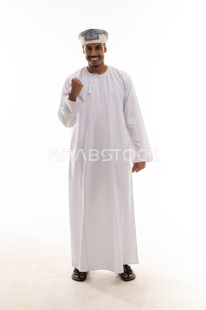 ضم قبضة اليد بقوة و رفعها للاعلى علامة على الفوز و التحدي , رجل عربي خليجي عماني يرتدي دشداشة بيضاء و عمامة , ايماءات السعادة و الابتسامة , الجسم بالطول الكامل , بورتريه خلفية بيضاء