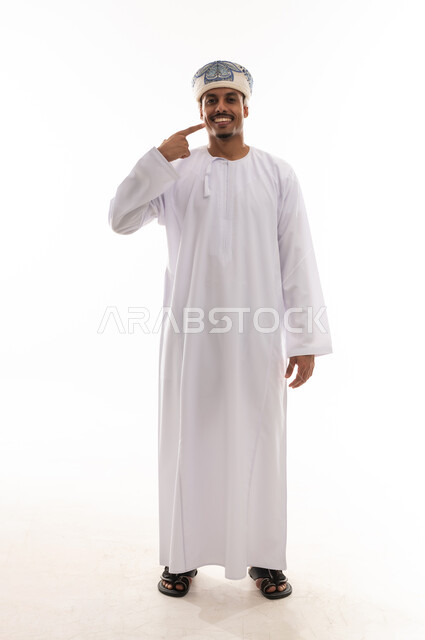 الاشارة الى الاسنان البيضاء الصحية باصبع السبابة , رجل عربي خليجي عماني يرتدي دشداشة بيضاء و عمامة يقف امام الكاميرا , تعابير الابتسامةو السعادة , بورتريه خلفية بيضاء , الجسم بالطول الكامل