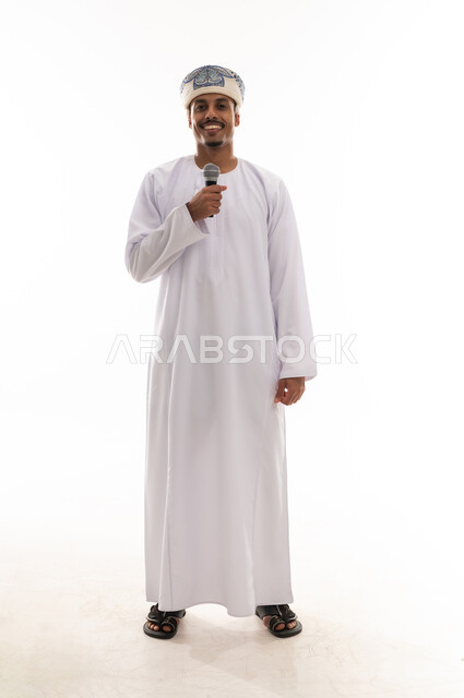 الوقوف امام الكاميرا و الابتسامة , رجل عربي خليجي عماني يرتدي دشداشة بيضاء و عمامة يحمل ميكروفون , الاستمتاع اثناء الغناء , تعابير السعادة , بورتريه خلفية بيضاء , الجسم بالطول الكامل