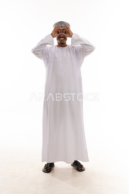 رجل عربي خليجي عماني يرتدي الدشداشة البيضاء و العمامة يقوم بعمل حركة دائرية باصبعي السبابة و الابهام اما العين اشارة الي المنظار المقرب , النظر بسعادة الي شئ ما , بورتريه الطول الكامل للجسم , خلفية بيضاء