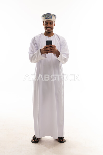 النظر بسعادة و الابتسامة الى الهاتف المحمول , رجل عربي خليجي عماني يرتدي دشداشة بيضاء و عمامة , الجسم بالطول الكامل , الوقوف امام الكاميرا , بورتريه خلفية بيضاء
