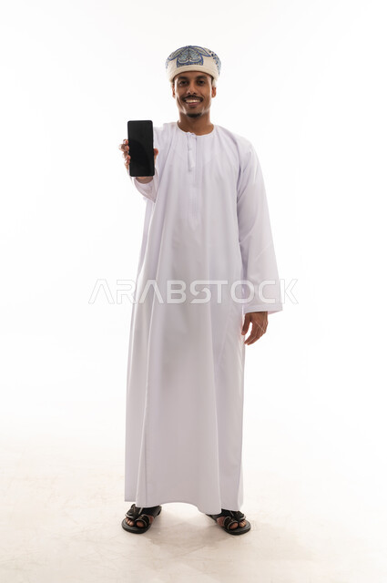 عرض شاشة هاتف محمول سوداء , رجل عربي خليجي عماني يرتدي الدشداشة البيضاء و العمامة يحمل الجوال امام الكاميرا , الجسم بالطول الكامل , بورتريه خلفية بيضاء , ايماءات السعادة و الفرحة