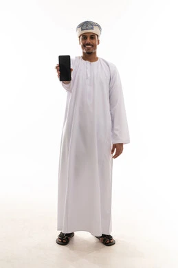 عرض شاشة هاتف محمول سوداء , رجل عربي خليجي عماني يرتدي الدشداشة البيضاء و العمامة يحمل الجوال امام الكاميرا , الجسم بالطول الكامل , بورتريه خلفية بيضاء , ايماءات السعادة و الفرحة