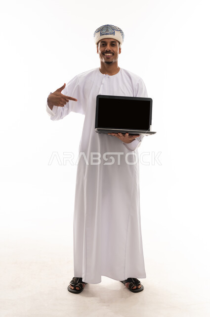 الاشارة الى الحاسوب المتنقل باصبع السبابة , رجل عربي خليجي عماني يرتدي الدشداشة البيضاء و العمامة يحمل جهاز الحاسب المتنقل , عرض شاشة سوداء فارغة امام الكاميرا , الجسم بالطول الكامل , بورتريه خلفية بيضاء