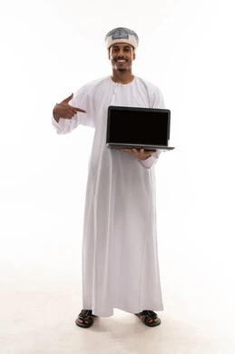 الاشارة الى الحاسوب المتنقل باصبع السبابة , رجل عربي خليجي عماني يرتدي الدشداشة البيضاء و العمامة يحمل جهاز الحاسب المتنقل , عرض شاشة سوداء فارغة امام الكاميرا , الجسم بالطول الكامل , بورتريه خلفية بيضاء