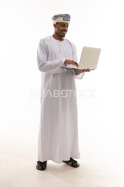 حمل الحاسب الالي المحمول الصغير بسهولة , رجل عربي خليجي عماني يرتدي الدشداشة البيضاء و العمامة يكتب على لوحة المفاتيح , ايماءات السعادة و الحماس , بورتريه خلفية بيضاء , الجسم بالطول الكامل