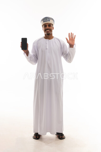 فتح قبضة اليد اشارة على الترجيب و السلام , رجل عربي خليجي عماني يرتدي دشداشة بيضاء و عمامة يعرض شاشاة هاتف محمول سوداء فارغة , بورتريه طول كامل للجسم , خلفية بيضاء
