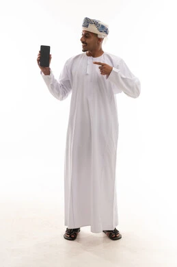 اشارة باصبع السبابة على الهاتف المحمول , رجل عربي خليجي عماني يرتدي الدشداشة البيضاء و العمامة , عرض شاشة سوداء فارغة , ايماءات السعادة و الابتسامة , خلفية بيضاء , بورتريه بالطول الكامل للجسم