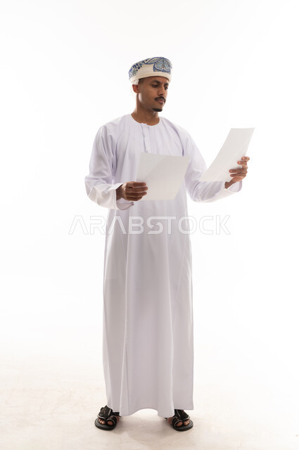 ايماءات التركيز و الجدية عند القراءة , رجل عربي خليجي عماني يرتدي دشداشة بيضاء و عمامة يحمل الاوراق , بورتريه الجسم بالطول الكامل , خلفية بيضاء , النظر الي الورقة
