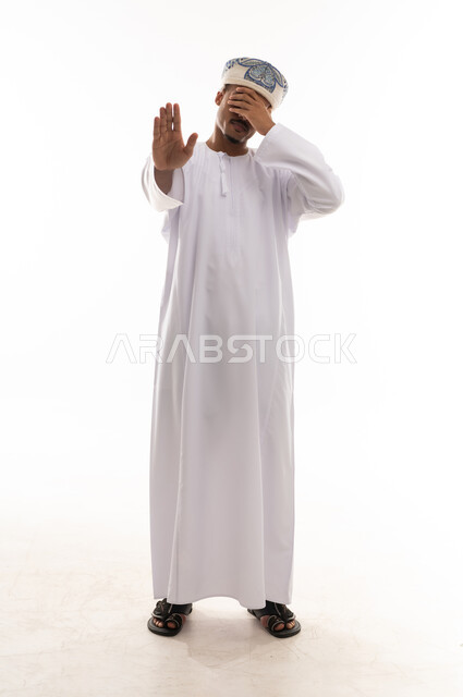 رفع باطن الكف اشارة على التوقف ووضع اليد الاخري على العين , رجل عربي خليجي عماني يرتدي الدشداشة البيضاء و العمامة يغطى عينيه لمنع الرؤية , بورتريه خلفية بيضاء , الجسم بالطول الكامل  , ايماءات الانزعاج